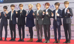 Eight men stand in a row, facing right and waving or holding peace signs