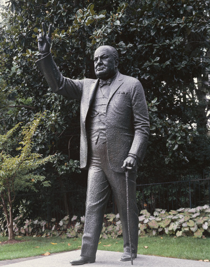 Statue on concrete surrounded by small lawn, flowers and screen of trees and shrubs