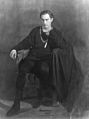Barrymore, cleanshaven, in an all-black costume as a brooding Hamlet, sitting on a chair, looking slightly to his right of the camera
