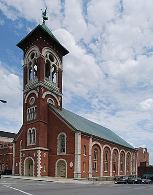 A brick church with elaborate stone decoration, greenish roofs and a tall square open tower at the front.