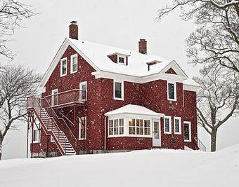 A two-story red house with a gabled roof pierced by two tall brick chimneys on the ends, and a large staircase going up the side, seen from its front left corner. Snow is falling and covering the ground