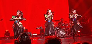 Three women wearing similar costumes consisting of a metallic top and dark skirt, performing on stage lit in bright red