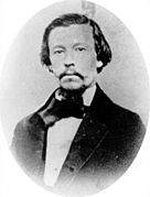 Man with facial hair and wearing a black suit and white shirt, facing camera