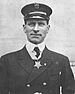 Head and shoulders of a man wearing a military jacket with a tie and high-collared white shirt underneath. A medal hangs from around his neck and his hat, a peaked cap, bears an anchor-shaped emblem with the letters "USN".