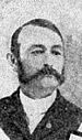 Portrait of a white man with a combover and sideburns connected to a mustache, wearing a dark suit.