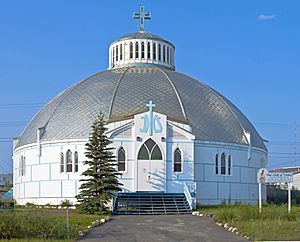 A one-story round building whose walls are white with large blue rectangles topped by a silvery dome with a blue cross and cupola at the center. From the camera a paved walk leads to blue wooden steps going up to its entrance, topped by wooden blue "IHS" letters. A aign in the yard at right says "Our Lady of Victory Church".