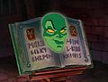 A green face, resembling that of a woman, is resting upon a book; the pages on either side are marked with Runic-like inscriptions. A cunning smile runs across her face, and her yellow eyes are looking upward.