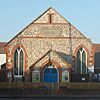 Front view of a tiny, plain chapel of irregular flintwork and red brick dressings. A dark stone plaque below a slit window near the roofline reads "WESLEYAN CHAPEL".  An entrance porch is flanked by two wide, low-set windows with pointed arches.