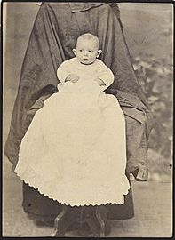 Baby in a long white dress on a chair draped plain dark fabric that may be two coats.