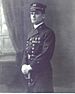 Gunner William Seach c. 1909 standing with a sword in his hands, wearing white gloves, a cap, and a long military coat with a row of medals on the left breast.