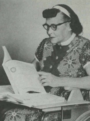 A white woman with dark hair, wearing a white headband, glasses and a patterned dress with short sleeves and a button front; she is seated in a wheelchair and reading a large softcover book