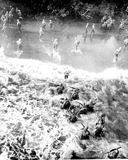 Heavily laden troops wade ashore through heavy surf.