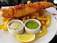 A large piece of battered fish on a plate of chips, served with ramekins of tartar sauce and mushy peas