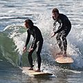 Two surfers wearing one-piece wetsuits, riding a wave.