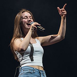 A young woman wearing a white tank top and jeans sings into a microphone she is holding with her right hand while smiling and raising up her other hand to gesture at the crowd.