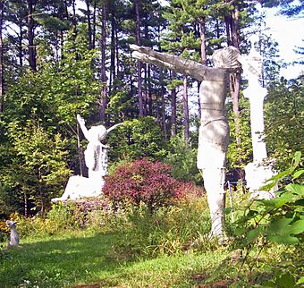 Four white sculptures sit in a grassy clearing in the woods, with a bush with dark pink flowers in the middle. Two are Native Americans wearing a single feather in bands at the rear of their head, holding a hand out and looking skyward. The one in the foreground stands while the one in the rear is seated. The other sculptures are a white column at the right rear and a baby unicorn and a giant frog at the left. There is a tall pine grove in the background.