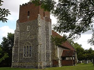 A church seen from the southwest; prominent is the west tower, its lower two stages in flint and the battlemented top stage in red brick; beyond it stretches the body of the church with its south porch