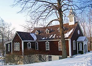 A dark brown wooden-sided building with a pointed roof, pointy-topped windows and a small pointed turret on the right end. Snow is on the surrounding ground and some of the roof, with a large bare tree in front. On the left side the light tan basement is visible.