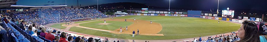 A night baseball game in progress in a small outdoor baseball stadium.