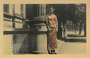A handcoloured photo of a woman wearing traditional Chinese clothings standing next to the State Library of Victoria, c. 1920s
