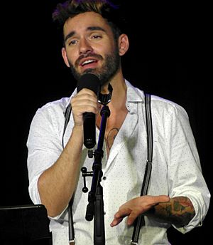 A brown-haired man with a beard and multiple tattoos performing on stage in a white shirt. He is holding a black microphone.