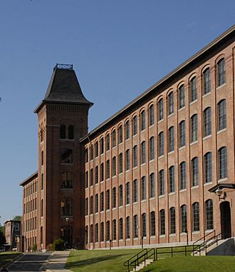 To the left are trees, and the background is a blue sky. From the far right, a building extends away to the left and into the distance, four storeys tall, each with an interrupted row of identical windows. At the middle of the building stands a tower with five storeys of windows topped by a square pyramidal roof with its tip truncated.