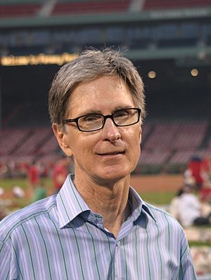 A portrait shot of a merry-looking middle-aged Caucasian male (John W. Henry) looking straight ahead. He has short greying hair, and is wearing a blue-striped shirt with the top button open. In the background is a baseball stadium.