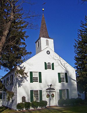 A white building with green shutters and pointed roof. At the front is a pointy tower with a round ball and weathervane on top