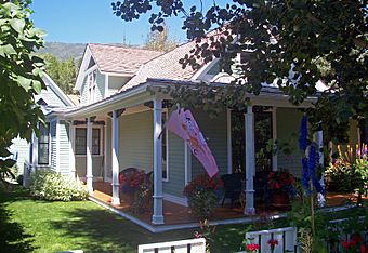 A one-story light blue house with a front porch supported by square pillars, seen from its front right, with some tree branches at the corners. There is a pink flag flying from the pillar closest to the camera. In the distance is a high ridgeline.