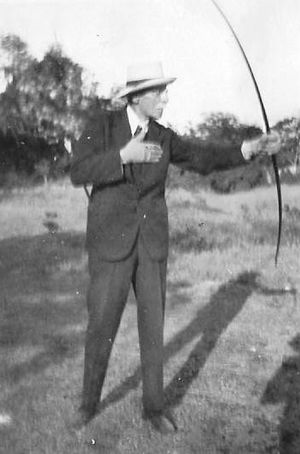 A black and white photograph showing an older man in a dark suit and tie, wearing a panama hat, with glasses and a light-coloured moustache. He is using a bow, his body is facing towards the camera, while he looks to the right, aiming the bow in the same direction. He is standing on grass, with shrubs and trees in the background.