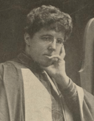 A white woman with short dark hair, head resting on one hand, wearing an academic robe