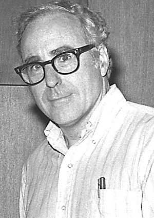 A middle-aged man with silver hair and glasses looks at the camera