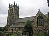 A sandstone church seen from the southeast; to the left is a tower with crocketted pinnacles and to the right is the body of the church with a clerestory and a gabled transept. In the foreground are gravestones and a bush