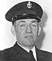 Head of a man wearing a dark jacket and tie and a peaked cap with an anchor and "USN" emblem on the front.