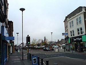 A road running off into the distance. On the right-hand side a three-storey building, with a shop front on the ground floor. Traffic lights and lamp standards are prominent. In the distance is the steeple of a church. The sky is grey.