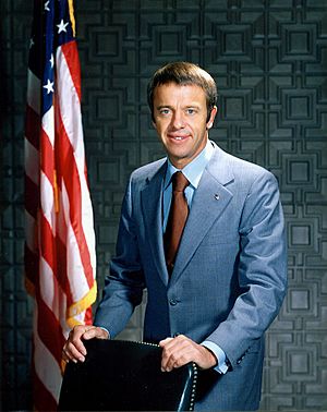 Shepard stands behind a chair wearing a blue suit. In the background is an American flag.