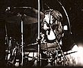 A black and white photograph of John Bonham wearing a headband and behind the cymbals of a drum kit