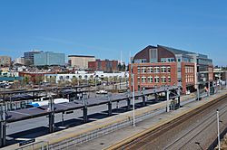 A train station with a three-story building and covered platforms seen against a background with several high-rise buildings in the distance.