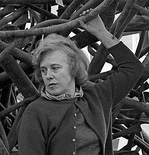 Claire Falkenstein, standing on one of her sculptures, her left arm raised above her head holding part of the sculpture