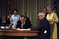 Two men at a desk with a document one is signing with their wives standing behind them