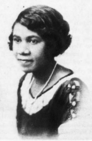 An African-American woman with wavy hair cut in a bob. She is wearing a necklace and a lace-collared top with a cut-out detail on the shoulder.