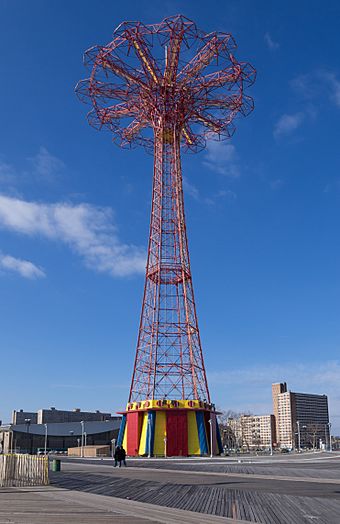The Parachute Jump, a tall red truss structure, and its pavilion, a red, yellow, and blue building near ground level. A wooden boardwalk can be seen in the foreground.