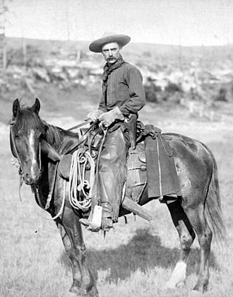 A black and white photograph of a cowboy posing on a horse with a lasso and rifle visible attached to the saddle