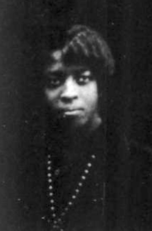 A young Black woman photographed mostly in shadows, wearing a dark dress with a strand of dark beads