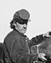 A white man with a thin mustache, wearing a military jacket and a forage cap low over his eyes. His side is turned to the camera and his is tending to a horse.