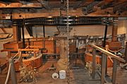 Interior view of the machinery on the first floor.  The "Great Spur Wheel" is an iron gear-wheel arranged horizontally on a vertical spindle, which spindle passes through the floor and is driven from below by the mill. on either side are millstones, set in a wooden drum-shaped housing.  They are driven by vertical iron shafts which, at the top, have a small gear mating with the great spur wheel.  The drive is thereby geared up to a much higher speed than the main vertical shaft from the wheel The floor is bare planks, the walls and apparatus are a golden coloured pale wood, and there is ancient whitewash on the walls.  The metal parts of the mechanism are painted a gloss black.