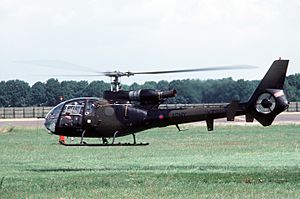 A small camouflaged helicopter hovers, just off the ground, over a field.