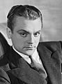 Black and white publicity photo of James Cagney—a white man with serious features and an arched eyebrow, dark eyes and hair combed back, wearing a suit and around 30 years of age—in the early 1930s.