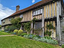 A view of the front of a Tudor-era cottage, with flowering plants climbing the weathered walls