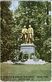 Picture postcard of a large bronze statue of two men on top of a stone pedestal. There are several wreaths that have been placed at the base of the pedestal. The statue is standing in a grassy clearing in front of trees. There is printing along the bottom of the postcard that says "Goethe and Schiller Monument, Wade Park, Cleveland, O."